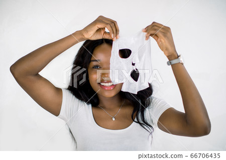 Face care and beauty treatments. Pretty smiling African woman holding a sheet moisturizing mask near her face, standing isolated on white background 66706435