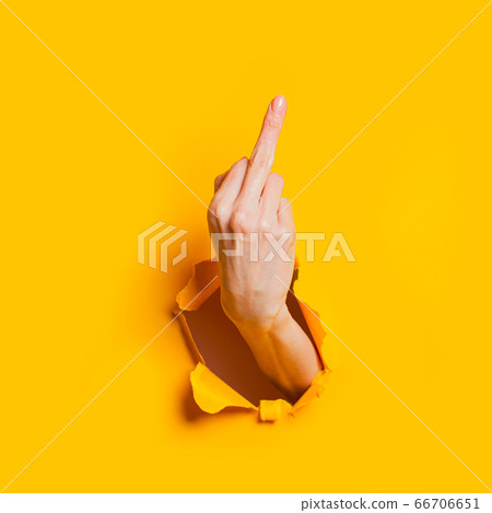 Female hand showing gesture meaning fuck you or fuck off through the hole in the yellow paper background. 66706651