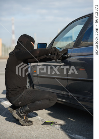 An offender in black clothes and mask is trying to steal a car in a parking lot. Stop criminality. 66712373