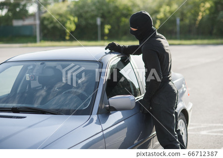 An offender in black clothes and mask is trying to steal a car in a parking lot. Stop criminality. 66712387