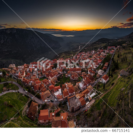 Aerial view of Delphi, Greece, the Gulf of Corinth, orange color of clouds, mountainside with layered hills beyond with rooftops in foreground Aerial view of Delphi, Greece, the Gulf of Corinth, orange color of clouds, mountainside with layered hills beyond with rooftops in foreground 66715141