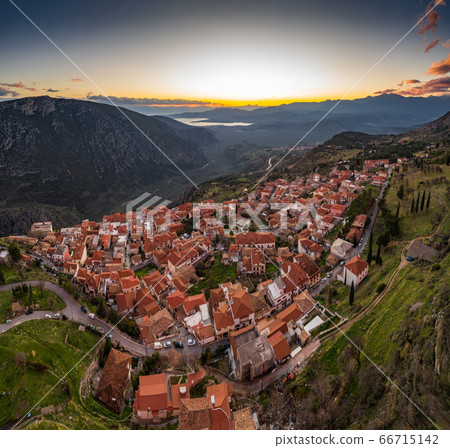 Aerial view of Delphi, Greece, the Gulf of Corinth, orange color of clouds, mountainside with layered hills beyond with rooftops in foreground Aerial view of Delphi, Greece, the Gulf of Corinth, orange color of clouds, mountainside with layered hills beyond with rooftops in foreground 66715142