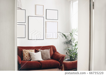 Blank picture frames mockups on white wall. White living room design. View of modern boho, scandi style interior with sofa, cushions, potted palm plant through open white door. Home staging concept. 66735220