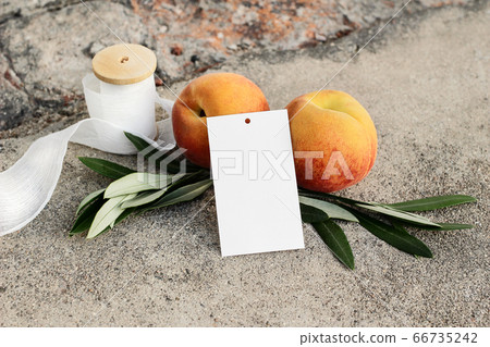 Summer wedding or birthday still life scene. Closeup of blank gift tag mock-up. Olive branches, silk ribbon and peaches fruit on grunge concrete background. Seasonal sale concept, no people. 66735242