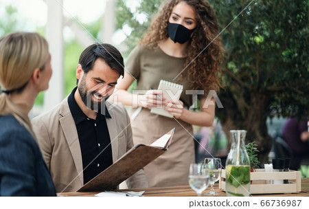 Waitress with face mask serving happy couple outdoors on terrace restaurant. 66736987