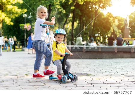 Two Cute adorable little caucasian siblings boy in helmet and girl having fun riding three-wheeled balance run bike scooter in city park or forest. Child first bike. Kids outdoor sport activities 66737707
