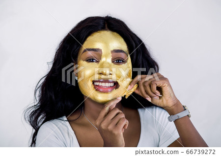 Skin care, beauty, spa. Pretty African woman with golden face mask. Close up of beautiful sexy girl taking off cosmetic peeling gold mask from healthy skin, posing to camera on white background Skin care, beauty, spa. Pretty African woman with golden face mask. Close up of beautiful sexy girl taking off cosmetic peeling gold mask from healthy skin, posing to camera on white background 66739272