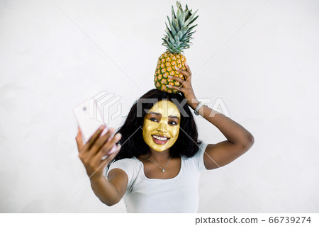 Attractive African girl with golden peel off mask, pineapple on her head, taking selfie photo with smartphone, enjoying relaxation and beauty treatment. Woman with facial mask making selfie Attractive African girl with golden peel off mask, pineapple on her head, taking selfie photo with smartphone, enjoying relaxation and beauty treatment. Woman with facial mask making selfie 66739274
