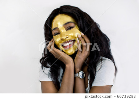 Spa, skin care, female facial treatment. Beauty close up portrait of joyful African woman with golden peel off facial mask touching her face and smiling to camera. Isolated on white background Spa, skin care, female facial treatment. Beauty close up portrait of joyful African woman with golden peel off facial mask touching her face and smiling to camera. Isolated on white background 66739288