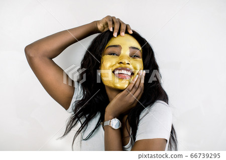 Cosmetic mask, beauty and skin care concept. Pretty charming African American lady with golden mask on her face, posing to camera with smile, enjoying her facial treatment Cosmetic mask, beauty and skin care concept. Pretty charming African American lady with golden mask on her face, posing to camera with smile, enjoying her facial treatment 66739295