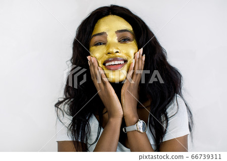 Pretty African woman with gold facial mask, touching her face with hands and looking at camera. Beautiful model with golden skin cosmetic touching her face. Beauty, skin care and treatment peeling Pretty African woman with gold facial mask, touching her face with hands and looking at camera. Beautiful model with golden skin cosmetic touching her face. Beauty, skin care and treatment peeling 66739311
