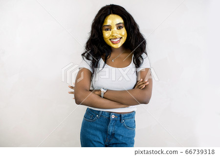 Portrait of young smiling African brunette woman in jeans and white t-shirt, standing on white background, with golden antiwrinkle mask on her face. Cosmetology and skin care concept. Portrait of young smiling African brunette woman in jeans and white t-shirt, standing on white background, with golden antiwrinkle mask on her face. Cosmetology and skin care concept. 66739318