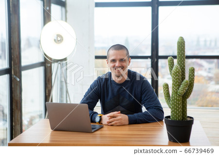 Smiling adult man looking in camera while sitting at desk with laptop. Smiling adult man looking in camera while sitting at desk with laptop. 66739469