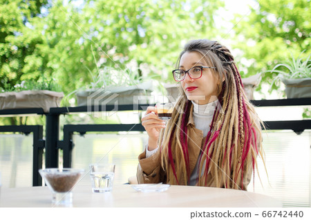 Young woman wearing glasses, casual nude trench and long dreadlocks is drinking coffee in outdoor cafe. Young woman wearing glasses, casual nude trench and long dreadlocks is drinking coffee in outdoor cafe. 66742440