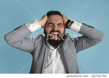 Millennial businessman holding his head and screaming in anger on blue background Millennial businessman holding his head and screaming in anger on blue background 66746740