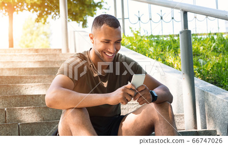 Happy black guy in sportswear relaxing after his workout on stairs and browsing internet on smartphone, outdoors Happy black guy in sportswear relaxing after his workout on stairs and browsing internet on smartphone, outdoors 66747026