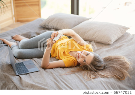 Cheerful young girl with laptop and mobile phone browsing net on bed at home Cheerful young girl with laptop and mobile phone browsing net on bed at home 66747031
