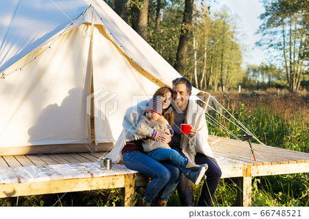 Happy family wraps blanket over themselves while sitting near canvas tent in the woods Happy family wraps blanket over themselves while sitting near canvas tent in the woods 66748521