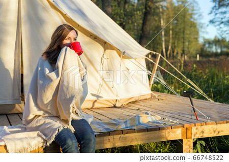 Young woman wraps blanket over herself while drinking coffee near canvas tent in the morning 66748522