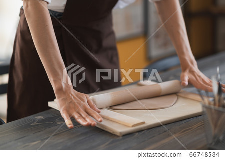Female hands flatting piece of clay lying on fabric using wooden rolling pin in ceramic studio. 66748584