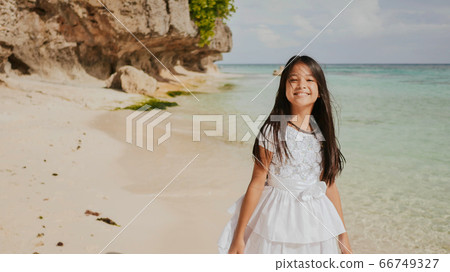 A charming and happy philippine teenage girl in a white summer dress is running along a tropical beach near the rocks. She is happily spinning. Childhood. Recreation. 66749327