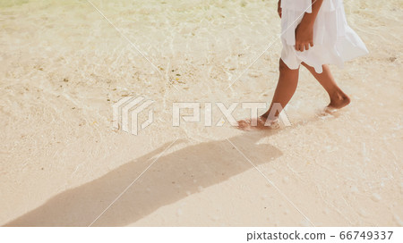 The naked and tanned legs of an philippine schoolgirl in a white dress walk on white sand, touching the foamy waves by the shore. Tropical landscape. Childhood. The naked and tanned legs of an philippine schoolgirl in a white dress walk on white sand, touching the foamy waves by the shore. Tropical landscape. Childhood. 66749337