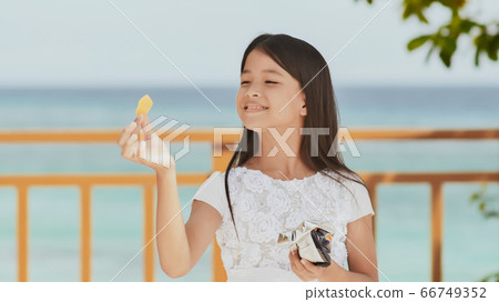 Philippine schoolgirl in white dress smiles, eating crispy potato chips. Tropical landscape. The palms. Summer. 66749352