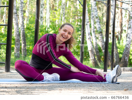 Smiling beautiful caucasian young woman in magenta sportswear does typical sitting legs stretching in sports area outdoor, in summer, selective focus. 66766436