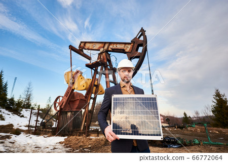 Businessman standing on an oilfield holding mini solar module next to an oil rig Businessman standing on an oilfield holding mini solar module next to an oil rig 66772064