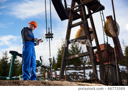 Oil engineer with a laptop standing next to an oil rig making notes in his computer 66772089