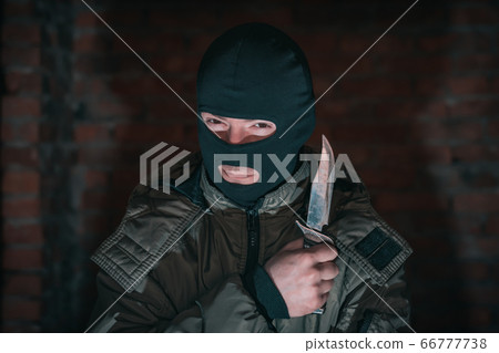 Suspicious look of a bandit in a balaclava with a knife on a background of a brick wall in the dark Suspicious look of a bandit in a balaclava with a knife on a background of a brick wall in the dark 66777738