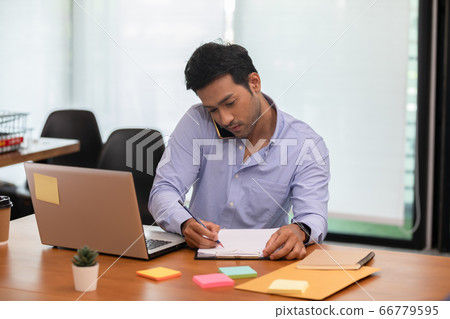 Asian indian business man talking with customer on mobile phone to get requirement and sale support holding pen in his hand at modern office or Co-Working Space,Startup Small Business Concept 66779595