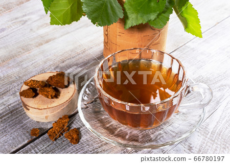 Glass figured cup of healing infusion of birch mushroom chaga, stump with chaga pieces and leaves on wooden table closeup. Chaga mushroom coffee or tea rich by antioxidants for immunity strengthening. 66780197