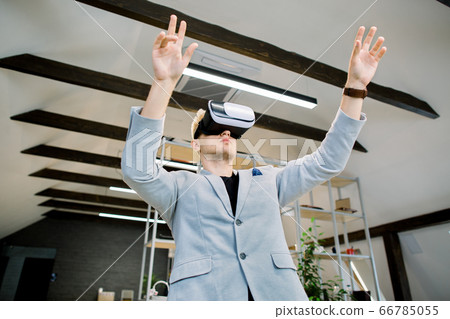 Young handsome creative businessman in blue jacket working in office with vr glasses on head. Man using VR headset for editing project or document in augmented reality Young handsome creative businessman in blue jacket working in office with vr glasses on head. Man using VR headset for editing project or document in augmented reality 66785055