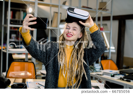 Beautiful girl with long dreadlocks, posing with virtual reality glasses on head, while making selfie photo on her smartphone, standing in modern office room. Hipster business woman wearing VR goggles Beautiful girl with long dreadlocks, posing with virtual reality glasses on head, while making selfie photo on her smartphone, standing in modern office room. Hipster business woman wearing VR goggles 66785056