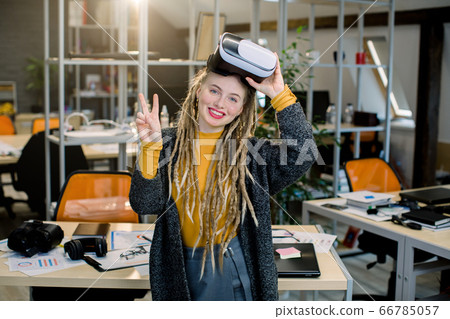 Happy smiling young blond girl with dreadlocks, posing with virtual reality goggles and gesturing victory sign, standing in modern office. VR glasses in office work. New technologies concept Happy smiling young blond girl with dreadlocks, posing with virtual reality goggles and gesturing victory sign, standing in modern office. VR glasses in office work. New technologies concept 66785057