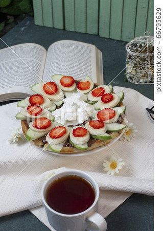 Waffles with banana and strawberry served with a cup of tea on a white tablecloth, a book around 66795629