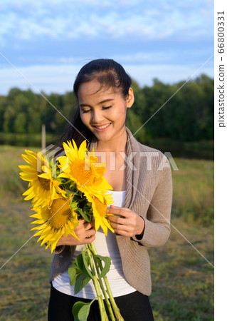 Young happy Asian woman holding bouquet of sunflowers against relaxing view of nature Young happy Asian woman holding bouquet of sunflowers against relaxing view of nature 66800331