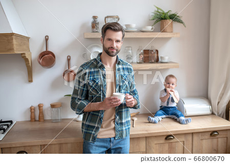 Young father standing with a cup in his hands, his little son sitting on the table Young father standing with a cup in his hands, his little son sitting on the table 66800670