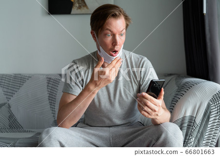 Young man with mask using phone and looking shocked at home under quarantine 66801663
