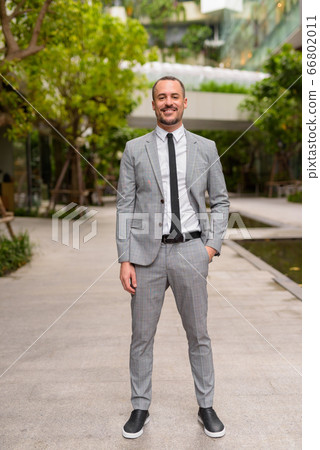 Full body shot of happy Hispanic bald bearded businessman smiling with nature in the city 66802011