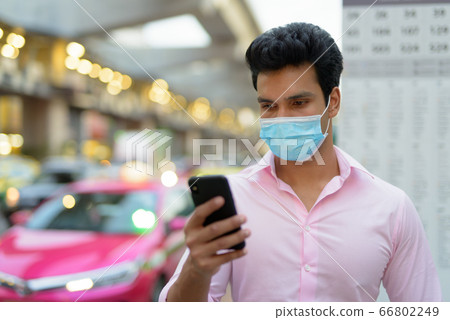 Face of young Indian businessman with mask using phone at the bus stop 66802249
