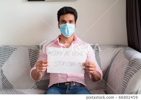 Young Indian businessman with mask showing Stay At Home sign while sitting in the living room under Young Indian businessman with mask showing Stay At Home sign while sitting in the living room under 66802459