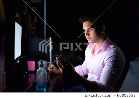 Young handsome Indian businessman using phone while working overtime at home during quarantine in Young handsome Indian businessman using phone while working overtime at home during quarantine in 66802582