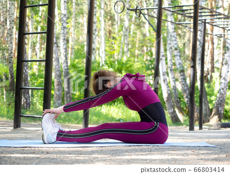 Fit caucasian young woman in magenta sportswear does seated forward fold in sports area outdoor, in summer, selective focus. 66803414