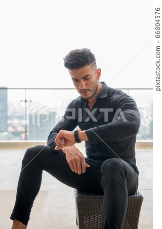 Young handsome bearded Persian businessman sitting and checking the time in the city Young handsome bearded Persian businessman sitting and checking the time in the city 66804576