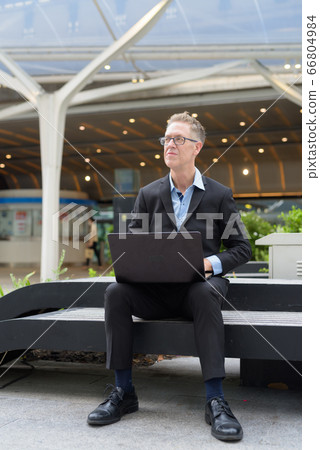 Mature businessman thinking and using laptop while sitting in the city outdoors 66804984