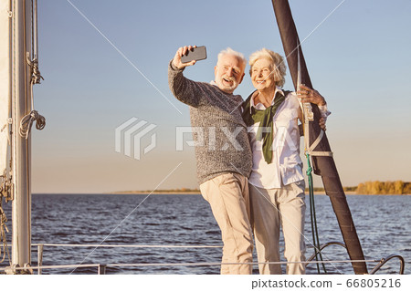 Happy senior couple standing on the side of sail boat or yacht deck floating in sea. Man and woman smiling, while taking selfie using smartphone 66805216
