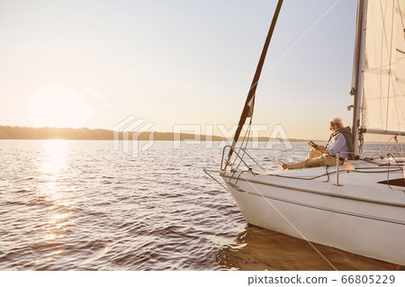 View of the sail boat or yacht floating in sea with relaxed senior man reading a book, sitting on the deck 66805229