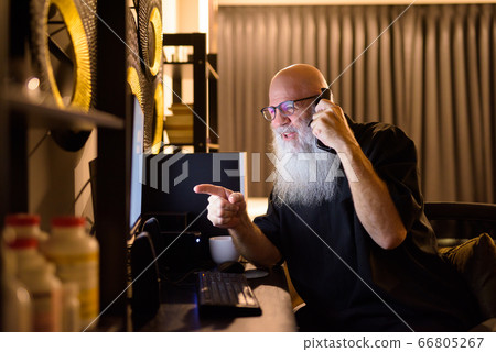 Happy mature bald bearded man talking on the phone while working overtime at home late at night Happy mature bald bearded man talking on the phone while working overtime at home late at night 66805267